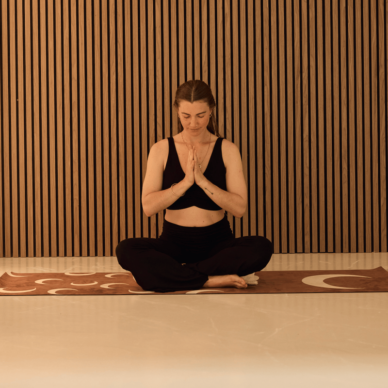 A woman in a black activewear set sits cross-legged on a brown yoga mat against a modern wooden slat wall, with her hands pressed together in a meditative pose.