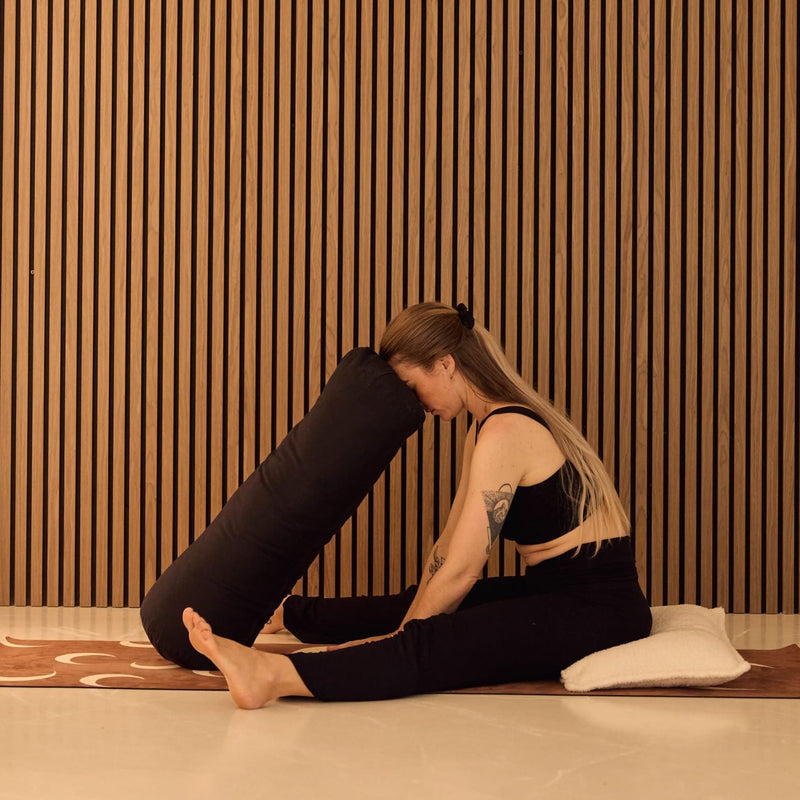 A woman in black activewear performs a yoga pose on a brown patterned mat, using a large black cylindrical bolster prop against her leg, with a white pillow behind her. She is positioned in front of a modern wall with vertical wooden slats.